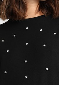 Svart tröja med rund halsringning, prydd med utspridda silverfärgade rhinestones över framsidan. Mjuk tyg med en texturerad yta.