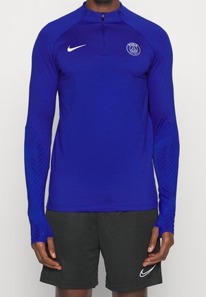 Mężczyzna w niebieskiej długorękawowej sportowej koszulce Paris Saint-Germain oraz czarnych szortach Nike z widocznymi logo na obu elementach odzieży.