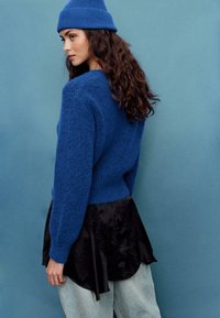 Modrý pletený sveter s textúrovanou tkaninou a rebrovanými manžetami, v kombinácii s čiernym saténovým viacvrstvovým topom a svetlými džínsami z denimu.