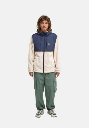 Junger Mann steht, trägt eine beige-navy Zip-Jacke, grüne Cargohosen und Turnschuhe, schaut mit neutralem Gesichtsausdruck nach vorne.