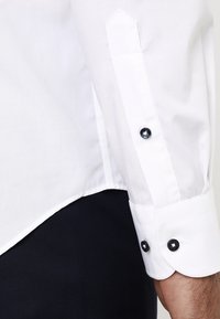 Polsino di una camicia bianca a maniche lunghe con bottoni neri indossata sopra pantaloni scuri, che mostra i dettagli dei bottoni e delle cuciture sul maniche.