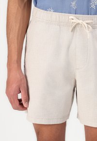 Beige, lightweight shorts with an elastic waistband and drawstring. Fabric has a slight texture, featuring side pockets and a clean hem.