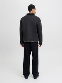 Schwarze Wollmischung Jacke mit Kragen und strukturiertem Finish, kombiniert mit locker sitzenden schwarzen Hosen, von hinten vor einem hellen Hintergrund betrachtet.