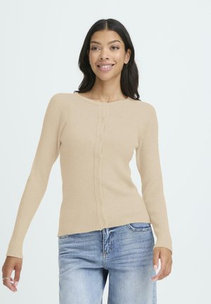 Junge Frau lächelt, trägt einen beigen geknöpften Langarmpullover und hellblaue Jeans vor einem schlichten hellen Hintergrund.