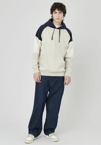 Sweatshirt à capuche en beige clair avec des accents navy, doté d'une poche frontale et d'un cordon de serrage. Porté avec un jean noir ample et des baskets.