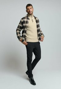 Herenoutfit met een zwart en beige geblokte flanellen shirt, beige geribde coltrui, zwarte slim-fit broek en zwarte enkelboots.