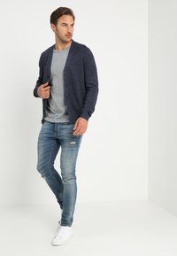 Marinblå cardigan, texturerad stickning, knappar framtill, bärs över en grå T-shirt. Ljust blå slitna jeans, vita sneakers, stående pose mot en neutral bakgrund.