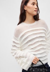 Pull rayé blanc en matière légère et texturée. Présente des couches horizontales avec des manches larges. Associe à un sac à main noir.