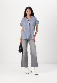 Striped short-sleeve button-up shirt in navy and white, paired with light gray flared jeans and white high-heeled ankle boots, holding a black bag.