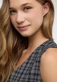 Primo piano di una persona con capelli castani lunghi e mossi, che indossa un indumento grigio a scacchi con scollatura a V, mostrando una pelle liscia e un trucco sottile.