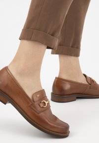 Mocassins en cuir marron avec un petit talon, présentant un détail de boucle dorée et une texture lisse, associés à un pantalon marron retroussé.