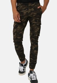Pantalons cargo camouflage en vert foncé et marron avec des ourlets à revers, de multiples poches latérales et une coupe ajustée, associés à des baskets noires.