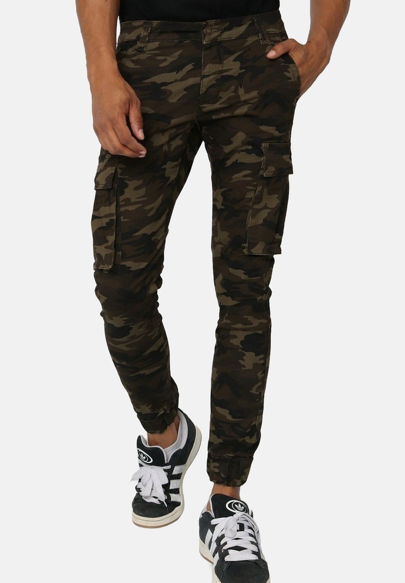 Pantalons cargo camouflage en vert foncé et marron avec des ourlets à revers, de multiples poches latérales et une coupe ajustée, associés à des baskets noires.
