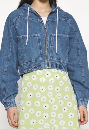 Kvinde iført en kort blå denim-hættetrøje med lynlås og store forlommer samt et lysegrønt skørt med hvidt blomsterprint.