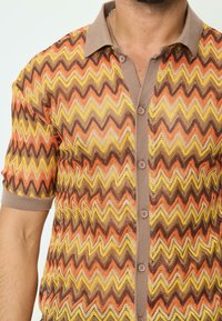 Chemise à manches courtes avec un motif en zigzag orange, jaune et brun. Comprend un col brun et des boutons sur le devant.