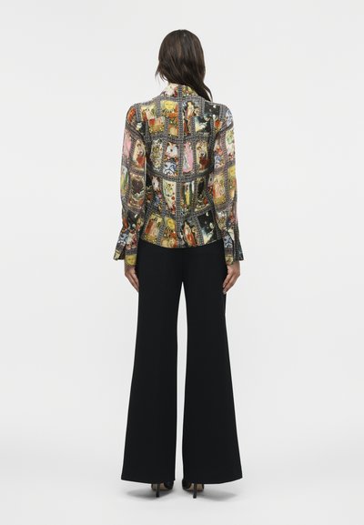 Γυναίκα με μακριά σκούρα μαλλιά που φοράει εφαρμοστή ζακέτα με πολύχρωμα σχέδια ταρώ και μαύρο παντελόνι φαρδιάς φάσας, εμφανιζόμενη από πίσω.