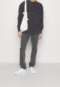 Grå sweatshirt med "TOMMY" tryckt i mörka, feta bokstäver, kombinerad med grå jeans och vita sneakers. En vit axelväska hänger över ena axeln.