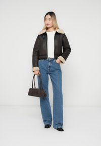 Chaqueta corta de cuero marrón con cuello de borreguito beige, camiseta blanca y jeans acampanados de un azul claro, sosteniendo un bolso oscuro y vistiendo zapatos negros.