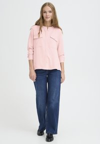 Chemise boutonnière rose pâle avec deux poches poitrine, associée à un jean bleu à jambes larges et des chaussures noires. Tissu doux avec une coupe décontractée.