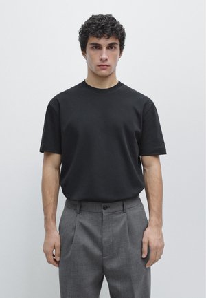 Giovane uomo con capelli ricci scuri che indossa una semplice maglietta nera a maniche corte e pantaloni grigi, in piedi davanti a uno sfondo grigio chiaro.