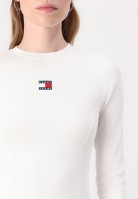 White ribbed long-sleeve shirt featuring a small black, red, and blue "TOMMY JEANS" logo patch at the center front. Smooth texture.