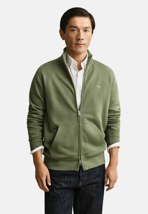 Man draagt een olijfgroen jack met ritssluiting over een wit overhemd en donkere spijkerbroek, staand tegen een effen witte achtergrond.