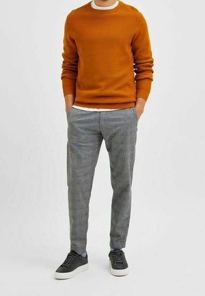 Oranje gebreide trui met ronde hals, gecombineerd met grijs geruite broek en zwarte sneakers. De trui heeft ribgebreide manchetten en zoom.