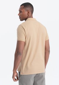 Hnědá polo košile vyrobená z texturované tkaniny, se stojáčkem a krátkými rukávy. Design má volný střih s bočními rozparky pro pohodlí.