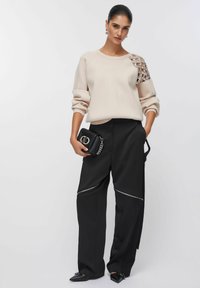 Bluză bej cu detaliu înnobilat pe umăr, asortată cu pantaloni negri largi cu fermoare argintii și o poșetă mică neagră.