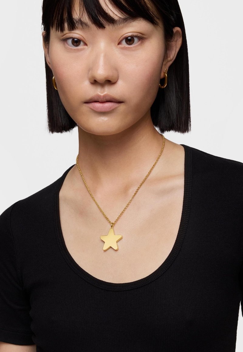 Collana con pendant a forma di stella dorata su una catena dorata, indossata su una maglietta nera a scollo rotondo. Finitura liscia e lucida, con un design semplice.