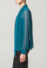 Blusa teal con maniche trasparenti e fluide e colletto rotondo. Presenta dettagli con bottoni sui polsini e un corpo dritto e aderente.