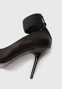 Cămașă neagră cu toc înalt, cu un superior din piele netedă, toc lucios și o curea la gleznă dintr-un material texturat negru cu literele brandului.