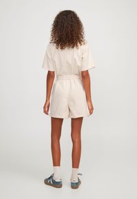 Monos corto de algodón beige con mangas cortas, cintura ceñida y pantalones cortos enrollados. El calzado incluye zapatillas azules con acentos blancos.