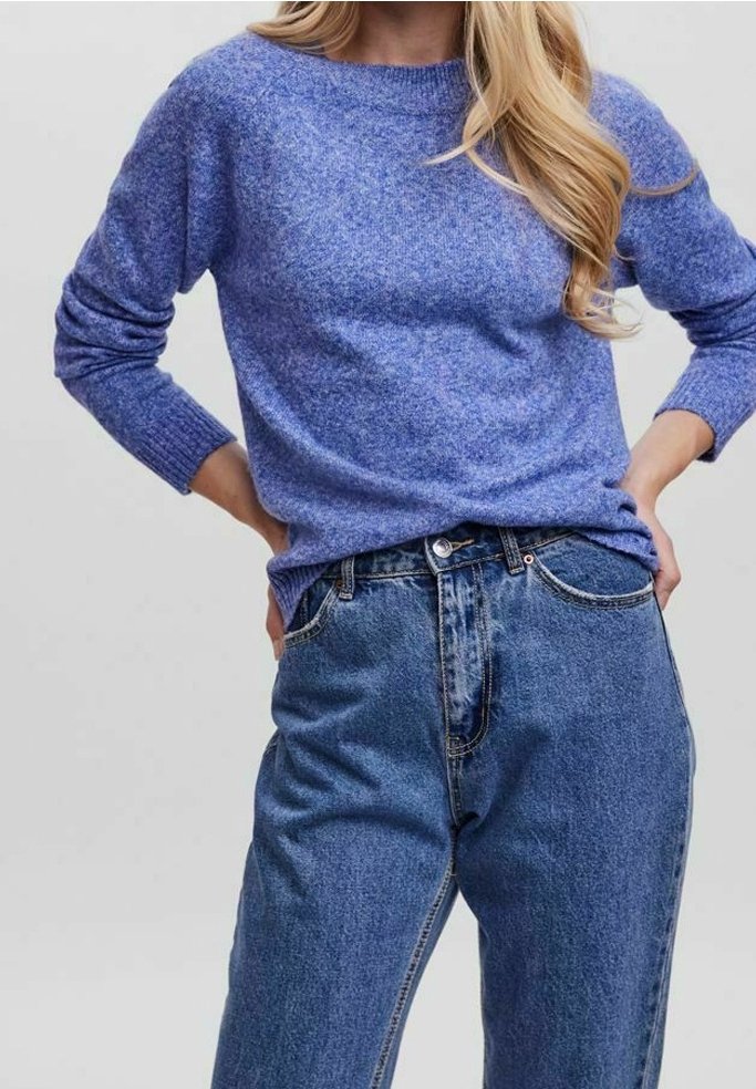 Femme aux longs cheveux blonds portant un pull en maille bleu rentré dans un jean bleu taille haute, mains posées sur les hanches.