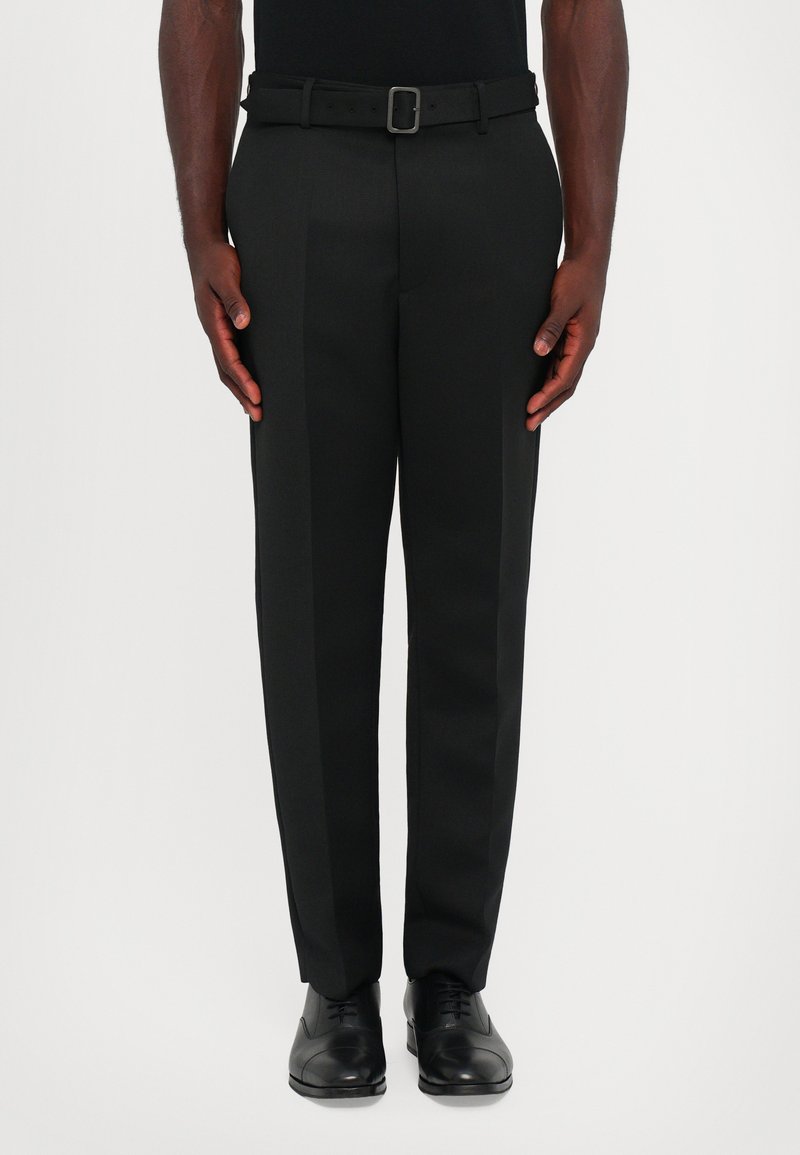 Pantaloni negri dintr-un material neted; au un croi drept și o curea elegantă cu o cataramă argintie. Purtați cu încălțăminte neagră.