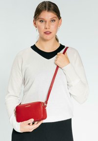 Rote Leder-Crossbody-Tasche mit strukturiertem Oberflächen, Reißverschluss und passendem Trageriemen. Das Model trägt einen weißen Pullover und schwarze Hosen.