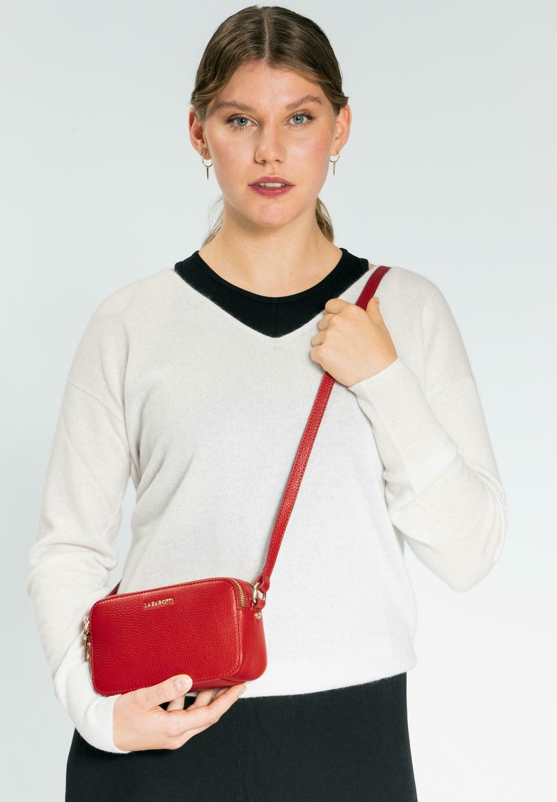 Rote Leder-Crossbody-Tasche mit strukturiertem Oberflächen, Reißverschluss und passendem Trageriemen. Das Model trägt einen weißen Pullover und schwarze Hosen.