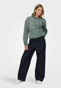 Oversized groene katoenen hoodie met een voorzak, gecombineerd met wijdvallende donkerblauwe jeans met zichtbare stiksels, gedragen met witte sneakers.