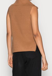 Pull sans manches marron côtelé avec un col montant, présentant une finition texturée et une coupe décontractée, porté avec un pantalon noir.
