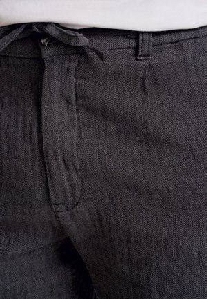 Mørkegrå hørfisk-mønstrede bukser med tekstureret stof, der har en bindebånd i taljen og en frontlomme. Subtile mønsterdetaljer er synlige.