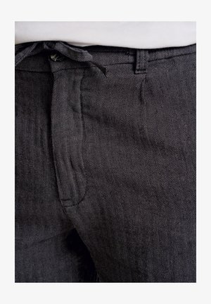 Pantaloni a spina di pesce grigio scuro in tessuto texturizzato, con vita a legaccio e tasca frontale. Dettagli del motivo sono visibili in modo sottile.