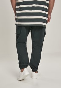 Donkergroene cargobroek met zijzakken, elastische boorden en een gladde textuur, gecombineerd met witte sneakers. Het model draagt een gestreept shirt.
