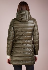 Mujer llevando una larga chaqueta acolchada verde oliva con capucha, vista desde atrás sobre un fondo beige liso.