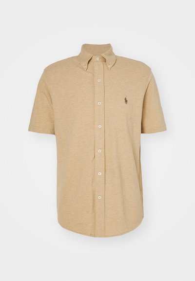 Camisa de manga curta em algodão bege, com colarinho abotoado, botões frontais e um pequeno logotipo no lado esquerdo do peito. Textura suave.