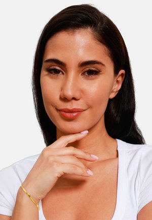 Modèle avec de longs cheveux noirs, portant un haut blanc, touchant délicatement son menton. Elle a un bracelet en or et un vernis à ongles rose clair.