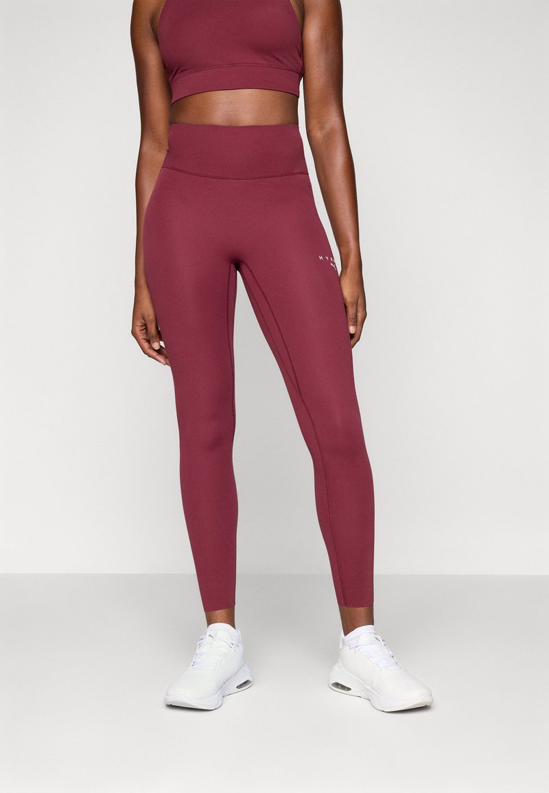Bordeaux-farbenes Sport-Bustier und Leggings-Set, das aus einem glatten Stoff, passgenauer Form und einem weißen Logo-Detail auf den Leggings besteht. Weiße Sportschuhe.