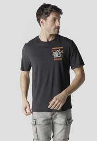 Man wearing black graphic t-shirt with tiger design and text, paired with light gray cargo shorts, standing against plain background.