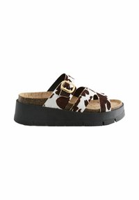 Sandalias de slide con estampado de vaca que presentan dos tiras anchas y una hebilla dorada. Suela de corcho sobre una plataforma de goma negra. Diseño plano para mayor comodidad.