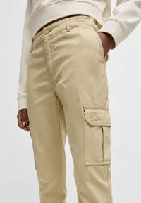 Pantaloni cargo beige con vestibilità rilassata, dotati di tasche laterali, una superficie liscia e un dettaglio del logo ricamato vicino alla tasca.