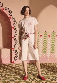 Biele krátke nohavice s pruhovaným látkovým opaskom, v kombinácii s ľahkým béžovým tričkom s malou farebnou grafikou. Červené topánky.